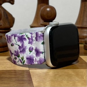 Może przedstawiać: Srebrny Apple Watch z czarnym ekranem i kwiatowym paskiem. Pasek ma białe tło z fioletowymi kwiatami i zielonymi liśćmi. Zegarek jest prezentowany na drewnianej powierzchni.