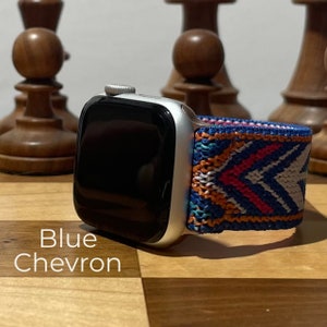 May include: A silver Apple Watch with a blue, orange, white, and pink patterned woven band. The band has a chevron pattern. The watch is on a wooden surface.
