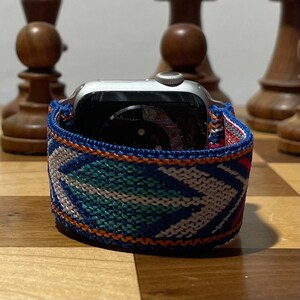 May include: A silver Apple Watch with a blue, orange, and white patterned woven band. The watch is on a wooden surface.