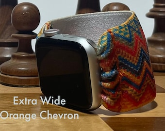 May include: A silver Apple Watch with a bright orange, blue, and yellow chevron patterned band. The band is extra wide and has a white backing. The watch face is black and the digital crown is visible. The watch is on a wooden surface.