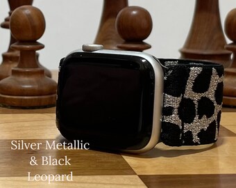 May include: A silver metallic and black leopard print Apple Watch band. The band is made of a soft, stretchy material and has a silver buckle.