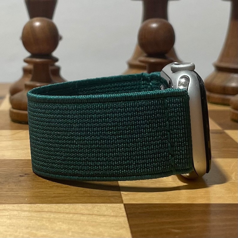 May include: A dark green sport band for an Apple Watch. The band is made of a soft, stretchy material and has a textured surface.