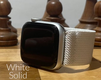 Könnte beinhalten: Ein weißes, durchgehendes Apple Watch Armband mit einer silbernen Apple Watch. Die Uhr liegt auf einer Holzoberfläche mit Schachfiguren im Hintergrund.