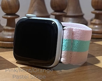 May include: A silver Apple Watch with a pink, mint and orange striped elastic band. The watch is on a wooden chessboard.