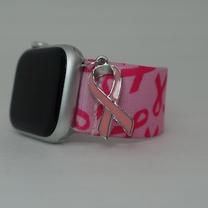 Könnte beinhalten: Eine silberne Apple Watch mit einem pink-weiß gemusterten Armband. Ein rosa Schleifenanhänger ist am Armband befestigt.