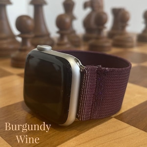 Könnte beinhalten: Eine silberne Apple Watch mit einem burgunderfarbenen elastischen Armband. Die Uhr liegt auf einer Holzoberfläche mit einem Schachbrettmuster.