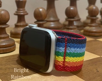 May include: A silver Apple Watch with a bright rainbow striped sport band. The watch is on a wooden chessboard.