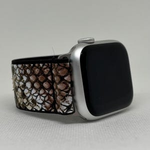 May include: Silver Apple Watch with a black screen and a snakeskin-patterned strap. The strap features a pattern of brown, tan, and white scales. The watch is angled, showing the strap and the watch face.