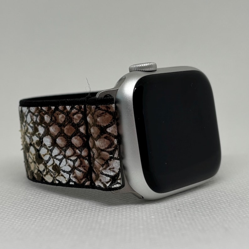 May include: Silver Apple Watch with a black screen and a snakeskin-patterned strap. The strap features a pattern of brown, tan, and white scales. The watch is angled, showing the strap and the watch face.