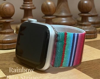 May include: A silver Apple Watch with a rainbow thin stripe elastic band. The watch is on a wooden surface.