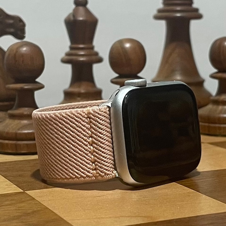 May include: A silver Apple Watch with a pink woven band. The watch is on a wooden chessboard.