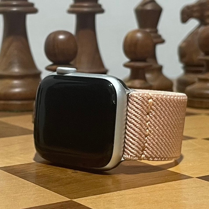 May include: A silver Apple Watch with a black face and a pink woven band. The watch is sitting on a wooden chessboard.