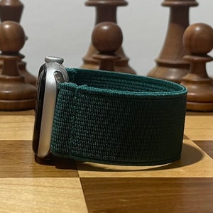 May include: A silver Apple Watch with a dark green woven nylon band. The watch is sitting on a wooden chessboard.
