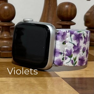 Może przedstawiać: Srebrny Apple Watch z czarnym ekranem i paskiem w fioletowe kwiaty. Pasek ma fioletowe kwiaty i zielone liście na białym tle. Słowo "Violets" jest wydrukowane w lewym dolnym rogu.