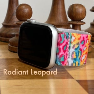 May include: A silver smartwatch with a black screen and a colorful Radiant Leopard band. The band features a vibrant leopard print in pink, yellow, and blue. The watch is displayed on a wooden surface.