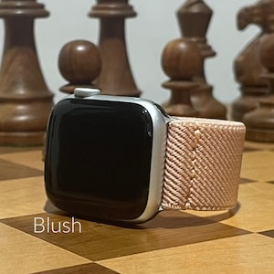 May include: A silver Apple Watch with a blush pink woven band. The watch is on a wooden surface with chess pieces in the background.