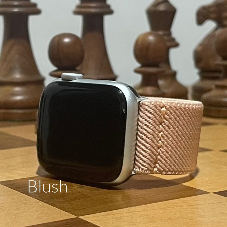 May include: A silver Apple Watch with a blush pink woven band. The watch is on a wooden surface with chess pieces in the background.