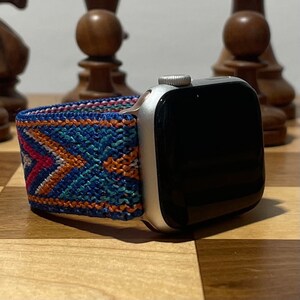 May include: A silver Apple Watch with a black screen and a colorful woven band. The band has a geometric pattern in shades of blue, orange, and pink.