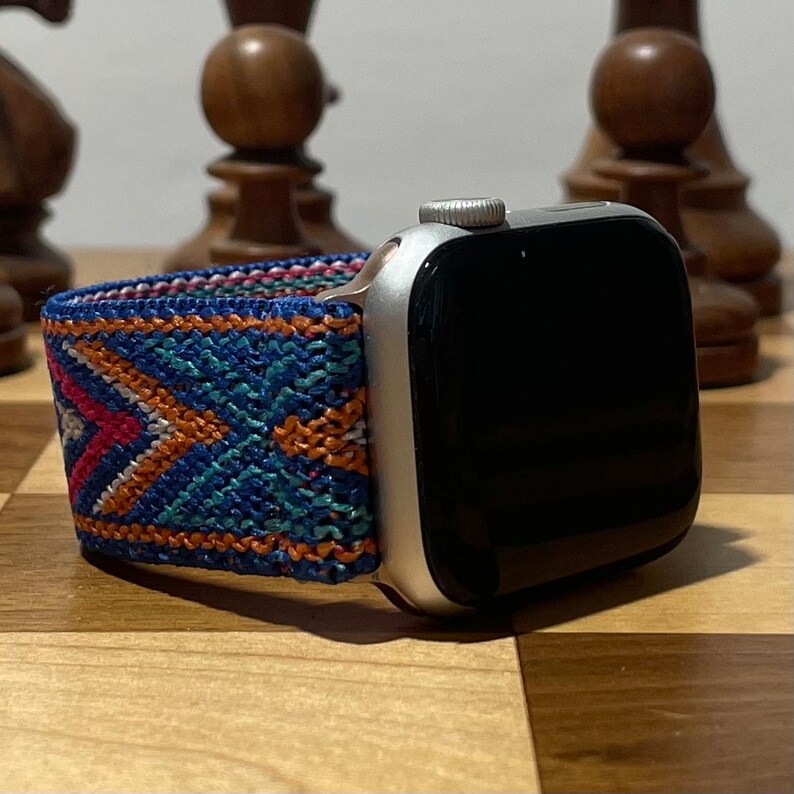May include: A silver Apple Watch with a black screen and a colorful woven band. The band has a geometric pattern in shades of blue, orange, and pink.