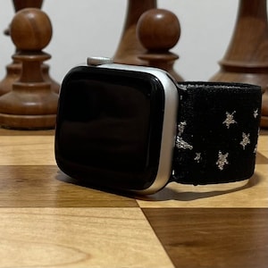 以下が含まれることがあります： 黒と銀の星柄のバンドが付いた銀色のApple Watch。時計は木製の表面の上に置かれています。