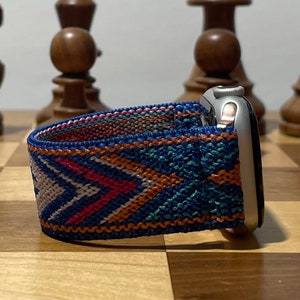 May include: A silver Apple Watch with a blue, orange, and white patterned woven band. The watch is on a wooden surface.