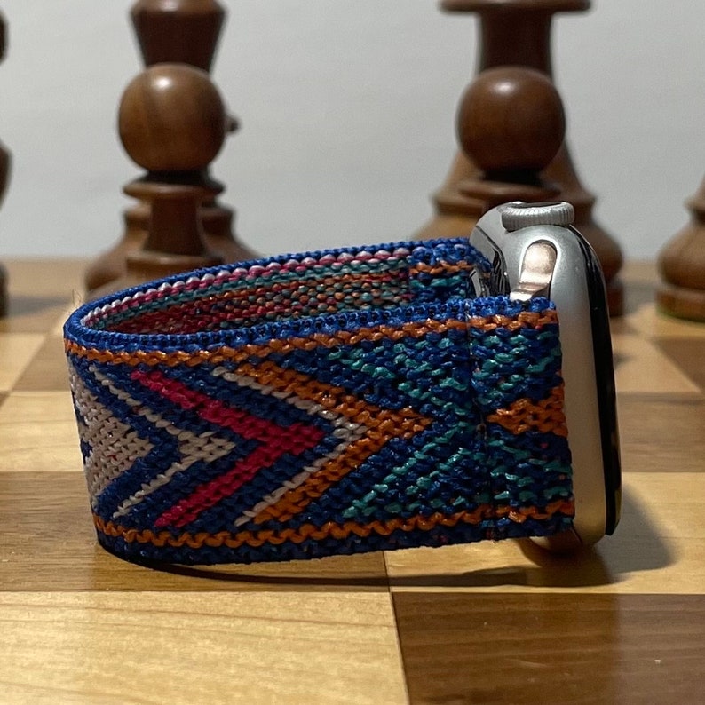 May include: A silver Apple Watch with a blue, orange, and white patterned woven band. The watch is on a wooden surface.
