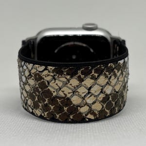 May include: A silver smartwatch with a snakeskin-patterned band in shades of brown, tan, and white. The band is wrapped around the watch face, showcasing the textured design. The band is approximately 2.5 cm wide.