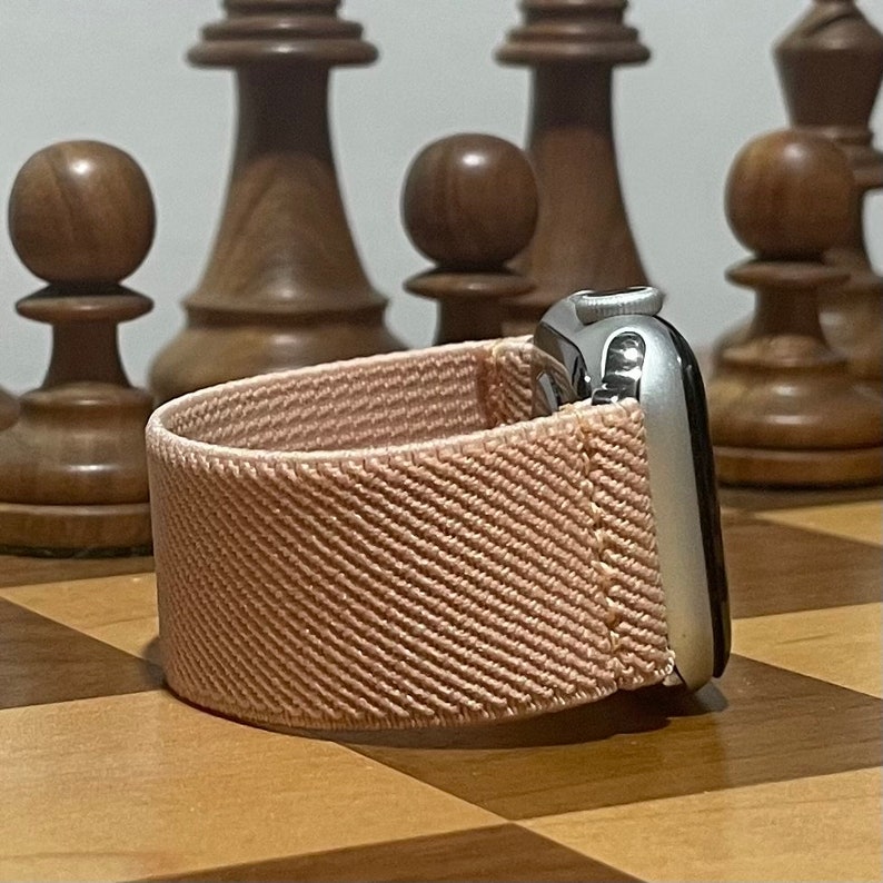 May include: A light pink woven Apple Watch band with a silver Apple Watch on a wooden chessboard.