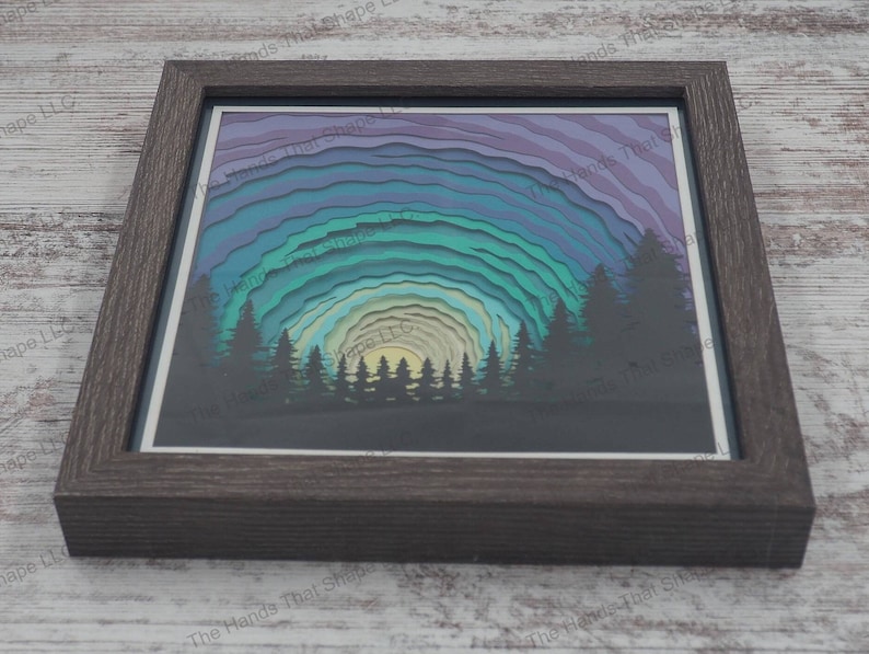 3D 'Northern Lights' shadowbox with a pine forest silhouette and colorful layers, in a glass-covered gray frame, made from 14 premium cardstock layers.