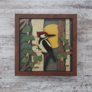 May include: A wooden wall art piece depicting a black and white woodpecker perched on a birch tree branch. The background features a yellow sun and green leaves. The artwork is framed in a dark brown wood frame.