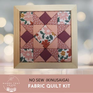 May include: A square wooden frame containing a no-sew Kinusaiga fabric quilt kit. The quilt features a geometric pattern of fabric squares and triangles in shades of pink, dark purple, and white with floral and polka dot prints.