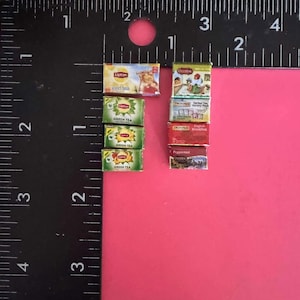 May include: A collection of miniature tea boxes, including Lipton iced tea, Lipton green tea, Twinings Herbal Tea Variety Pack, Twinings English Breakfast, and Celestial Seasonings Peppermint. The boxes are arranged in a vertical stack on a pink surface.