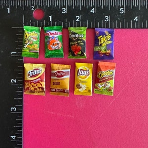 May include: Eight miniature bags of popular snack brands, including Cheetos, Fritos, Doritos, Lay's, and Takis. The bags are arranged in two rows of four on a pink surface.