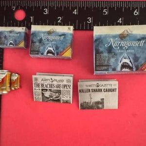 May include: Miniature Narragansett lager beer boxes, two small beer cans, and two Amity Island newspapers with headlines reading The Beaches Are Open and Killer Shark Caught, arranged next to a black ruler showing inch measurements.