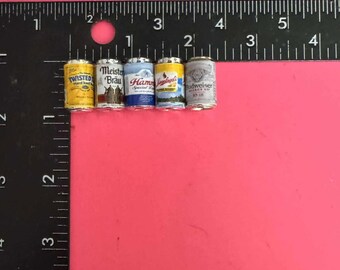 May include: Miniature beer cans, including Twisted, Meister Brau, Hamm's, and Budweiser, are arranged on a pink surface next to a ruler. The cans are small, with detailed labels and a metallic finish, showcasing a variety of brands.