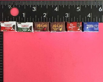 May include: A row of seven miniature coffee boxes, including Dunkin' Donuts, McCafe, Tim Hortons, and Maxwell House. The boxes are arranged in a line on a pink surface with a ruler behind them.
