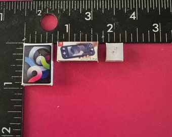 May include: Three miniature boxes, one with a blue Nintendo Switch Lite game console, one with a colorful abstract design, and one with a white surface. The boxes are on a pink surface with a black ruler in the background.