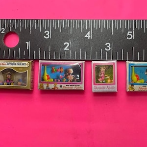 May include: Four miniature toy boxes with colorful illustrations of dolls and toys. The boxes are labeled "Kiddie's Kare Antique Doll Set" and "Steiff Kiddie".