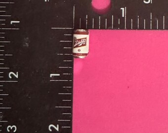 May include: A miniature aluminum can with a red and white label that says "Schlitz" on it. The can is about 1 inch tall and is sitting on a pink surface.