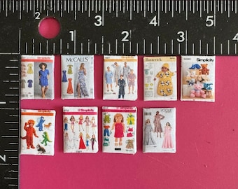 May include: A collection of eight miniature sewing patterns, including Simplicity, McCall's, Vogue, and Butterick brands. The patterns feature a variety of designs, including dresses, pajamas, and stuffed animals.