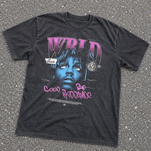 May include: A charcoal gray short-sleeve t-shirt featuring a graphic print of a person's face in blue tones. The text WRLD, Juice, and Good Bye & Good Riddance are printed in pink and white. The shirt has a crew neckline and a vintage-style wash.