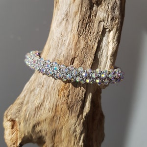 Może przedstawiać: Bransoletka z koralików srebrnych i tęczowych kryształów. Bransoletka wykonana jest z małych, fasetowanych koralików, które są nawleczone na sznurek w ciasnym wzorze. Bransoletka jest pokazana na kawałku drewna unoszącego się na wodzie.