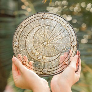 Può includere: Vetrata rotonda con un design celeste. Presenta una luna crescente, stelle e un motivo centrale a stella, il tutto incorniciato da un bordo in metallo scuro. Gioielli in vetro trasparente sono incastonati sul bordo.