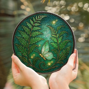 May include: A round, stained-glass art piece featuring a butterfly and fern design in shades of green and gold. The butterfly has a light green body and gold accents. The artwork is held by hands, with a chain for hanging.