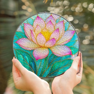 May include: A round stained glass piece featuring a lotus flower design. The flower has pink and yellow petals with green leaves. The glass is held by a black metal frame and chain.
