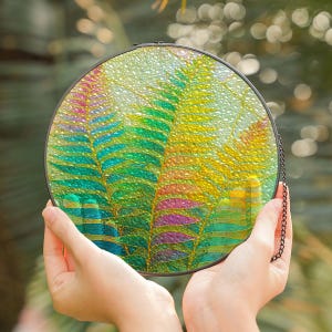 Puede incluir: Un panel redondo de vidriera con un diseño de helecho en tonos verdes, amarillos, azules y morados. El vidrio está sujeto en un marco de metal negro con una cadena para colgar. El panel se sostiene frente a un fondo verde borroso.