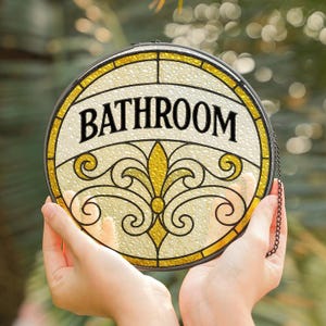 May include: Round stained glass bathroom sign with a black chain. The sign features the word "BATHROOM" in black letters. The design includes yellow and gold accents with a decorative floral pattern. The sign is held by two hands.