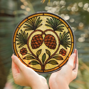 Könnte beinhalten: Ein rundes Buntglasfenster mit einem von der Natur inspirierten Design. Das Kunstwerk zeigt zwei braune Tannenzapfen, grüne Blätter und orangefarbene Blüten auf gelbem Hintergrund. Das Fenster ist mit einem dunklen Metallrahmen und einer Kette zum Aufhängen versehen.