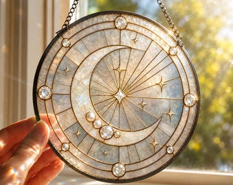 May include: A circular suncatcher featuring a crescent moon and star design with iridescent, textured glass panels held by a dark metal frame. The piece is accented with small clear crystals and pearls, suspended by a thin metal chain.