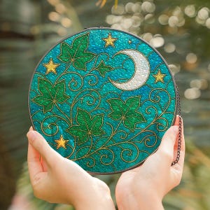 以下が含まれることがあります： ターコイズブルーの背景に、緑のツタの葉、金の星、白い三日月が描かれた円形のステンドグラスアート。 作品は手で持たれており、吊り下げ用の黒いチェーンが付いています。